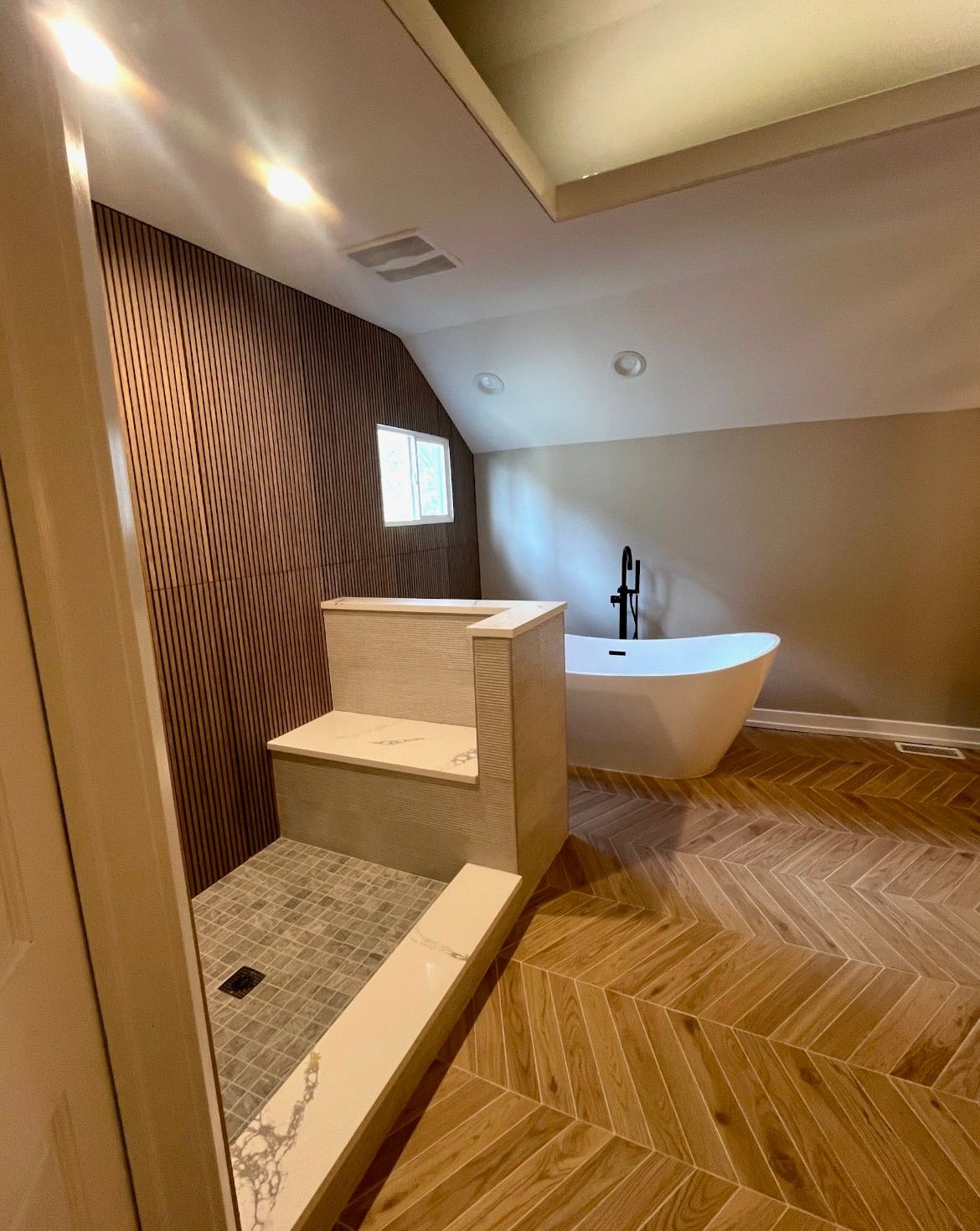 Modern bathroom with freestanding tub, wood panel walls, and herringbone pattern wooden floor.